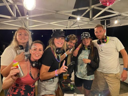 A group of six people wearing headphones, smiling and posing under a tent at night.
