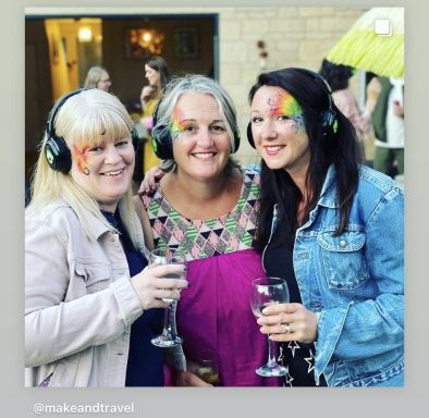 Three women smiling and holding glasses, with colourful face paint at a gathering.