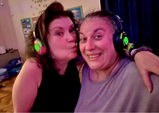 Two women smiling and wearing headphones, enjoying a playful moment together.